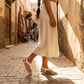 White bohemian flat t-strap sandals adorned with 3D floral appliques and crystal accents, worn by a model with an elastic slingback strap during a stroll in a sunlit cobblestone street.