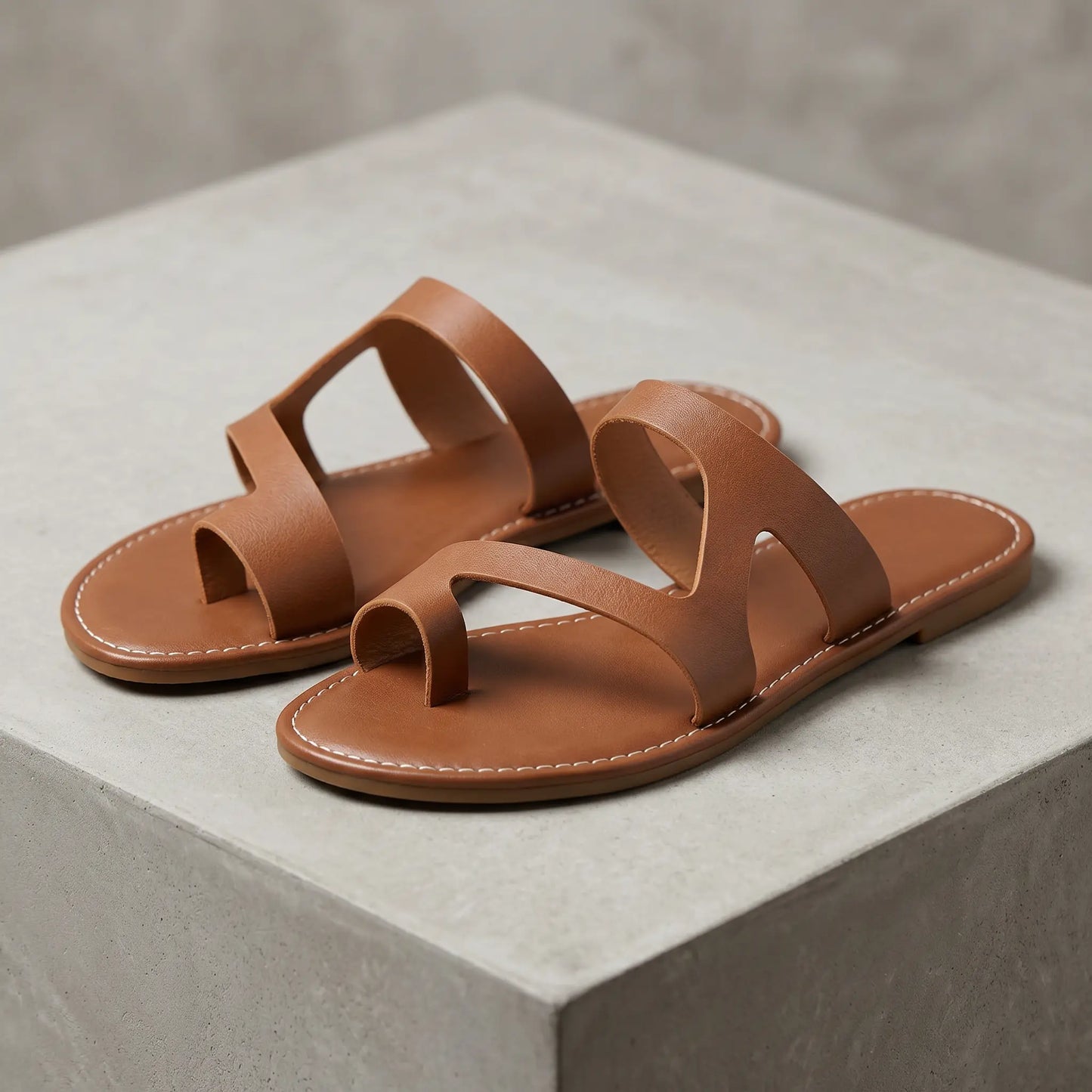 Elegant brown faux leather flat sandals with a toe loop and asymmetrical strap design, detailed with white contrast stitching around the flat sole. Shown from a three-quarter view.