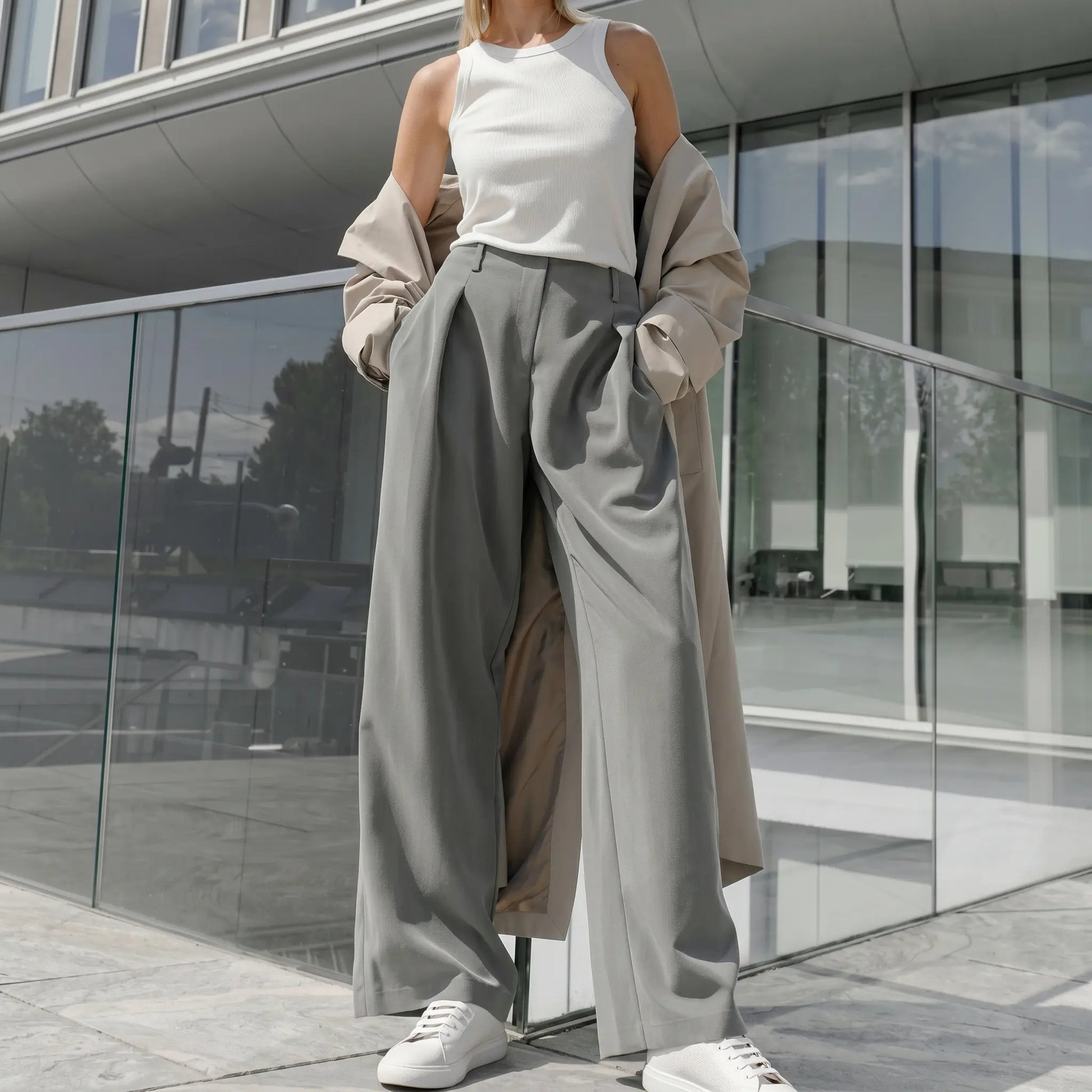 Woman wearing elegant grey wide-leg trousers with sharp front pleats and a fluid drape, styled with a white tank top and beige trench coat.