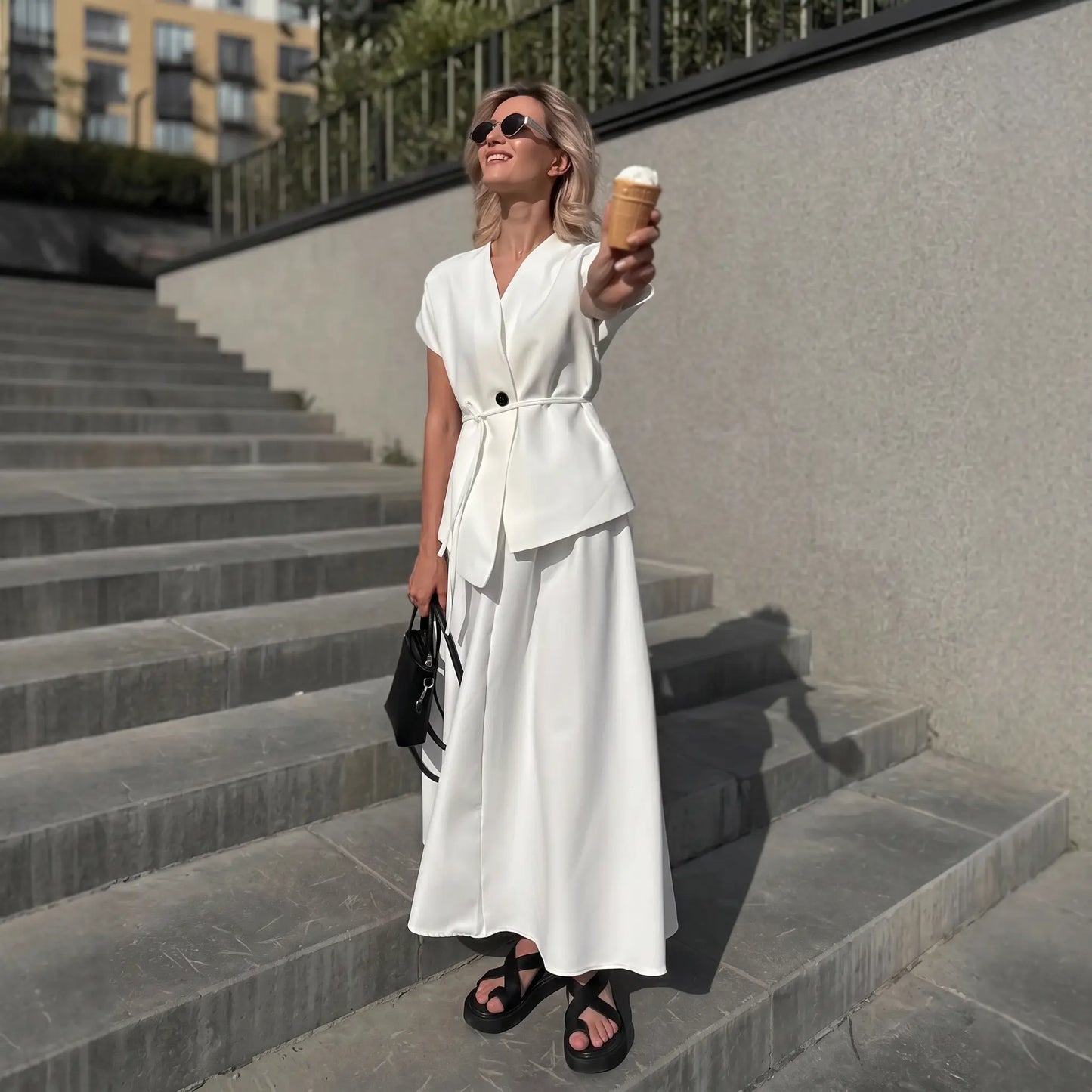 Woman wearing an elegant white two-piece set featuring a sleeveless V-neck top with a tie belt and an A-line midi skirt, standing on outdoor stairs while holding an ice cream cone.