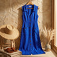Elivora blue linen midi dress with button-down front and pockets, displayed on a hanger against a stone wall with a straw hat and pampas grass.