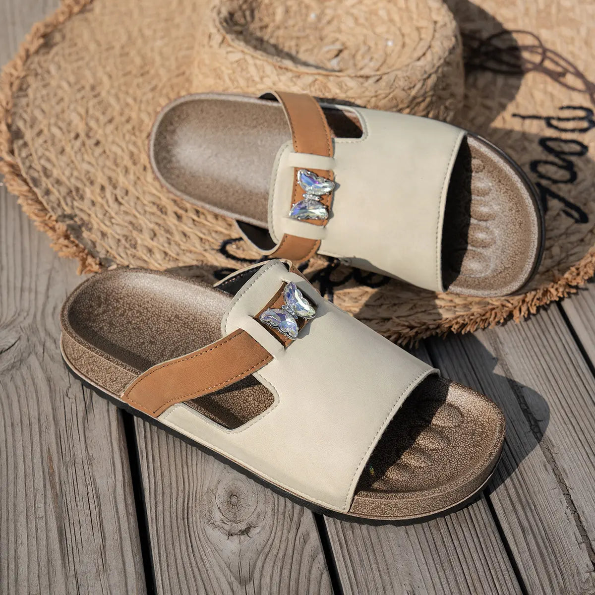 Cream embellished flat slide sandals with tan accent straps and clear butterfly details, styled on a straw hat.