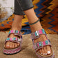 Embellished platform slide sandals with rhinestones and colorful stones, featuring double buckle straps and a cork-effect footbed, shown on a model's feet with grey jeans. A boho chic accessory for summer.