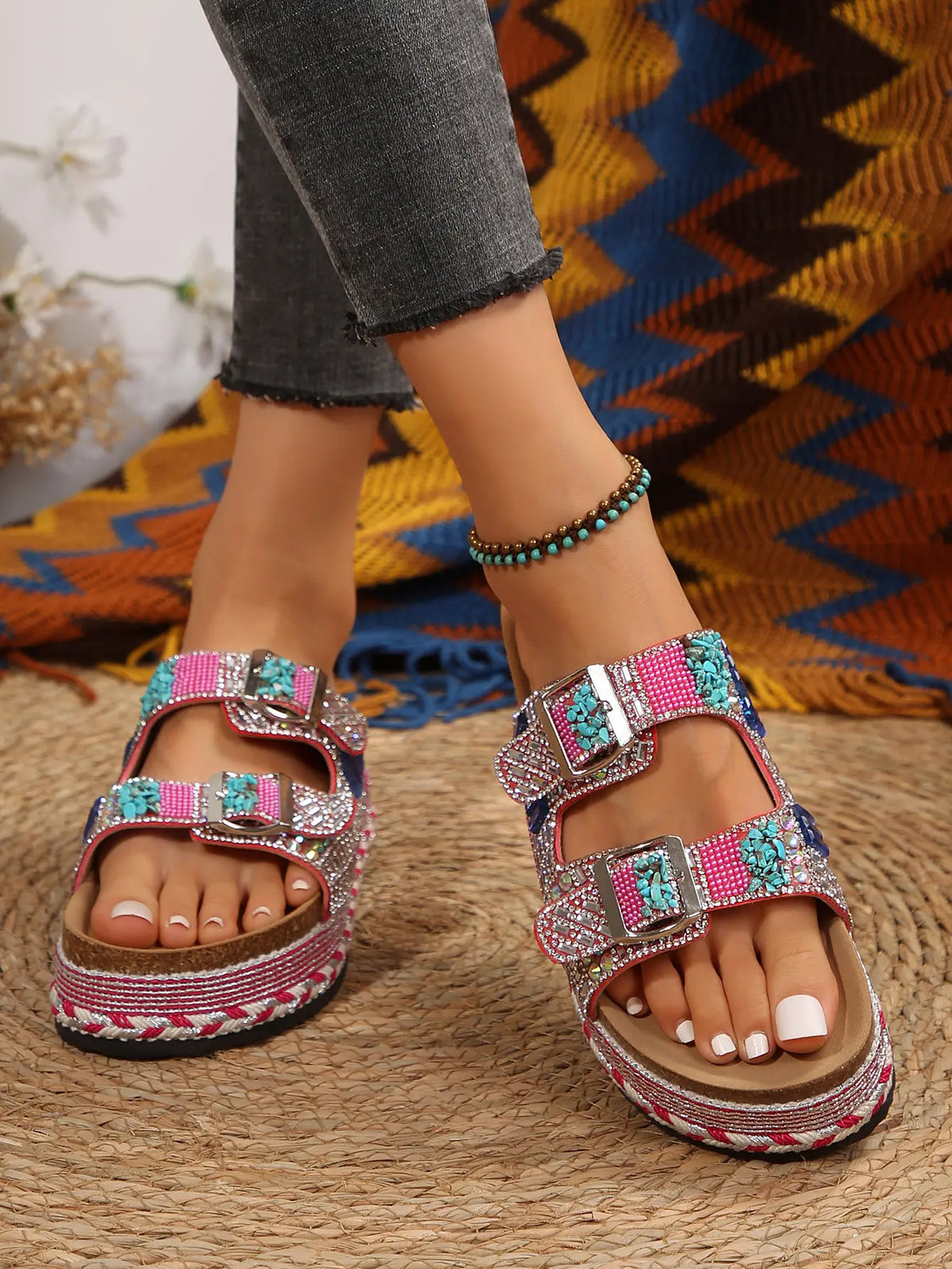 Embellished platform slide sandals with rhinestones and colorful stones, featuring double buckle straps and a cork-effect footbed, shown on a model's feet with grey jeans. A boho chic accessory for summer.