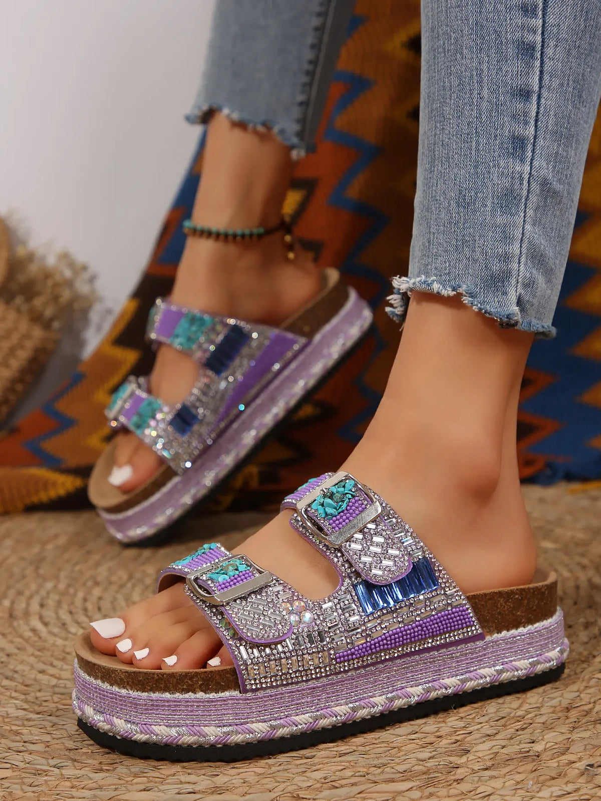 Woman's feet wearing embellished platform slide sandals with purple straps, rhinestones, beads, and blue resin stones, paired with denim jeans.