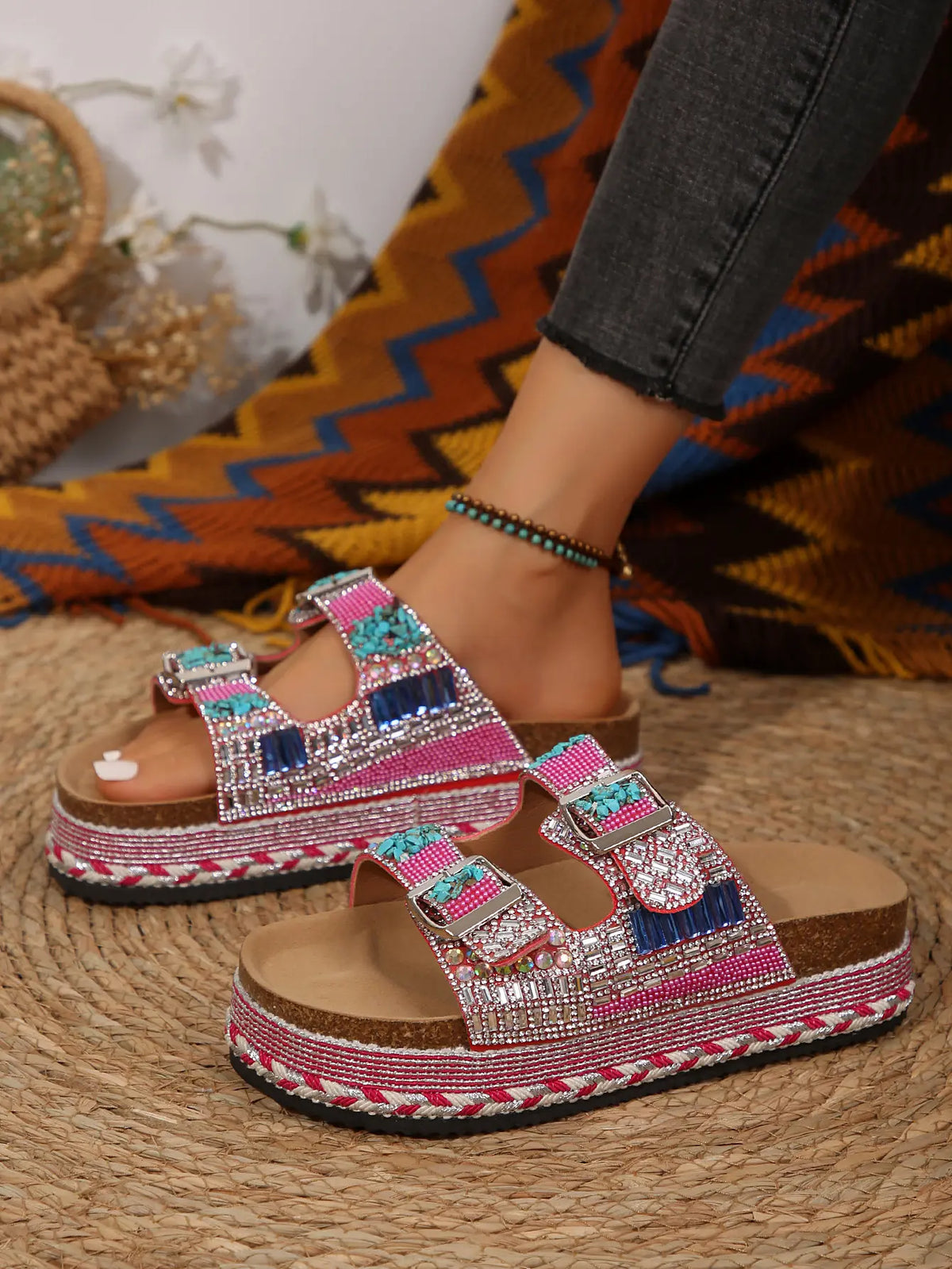 Embellished platform slide sandals with double buckle straps, adorned with dazzling rhinestones, pink beads, and turquoise resin stones, worn on a foot with a bohemian backdrop.