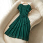 Emerald green A-line midi dress with sheer cap sleeves, a classic round neckline, and a self-tie bow cinching the waist. The fluid, double-layered asymmetric skirt cascades gracefully over a cream velvet armchair.