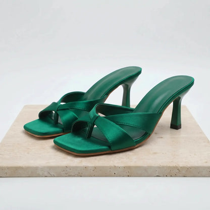 Emerald green satin square-toe stiletto mules with a knotted strap detail and a flared heel, displayed on a marble slab against a white background.
