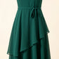 Close-up of an emerald green A-line midi dress, showcasing the self-tie fabric sash at the cinched waist and the fluid, asymmetric double-layer chiffon skirt with a cascading hem.
