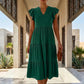 Model wears an emerald green tiered midi dress with a V-neck and ruffle cap sleeves, walking in a modern architectural hallway.