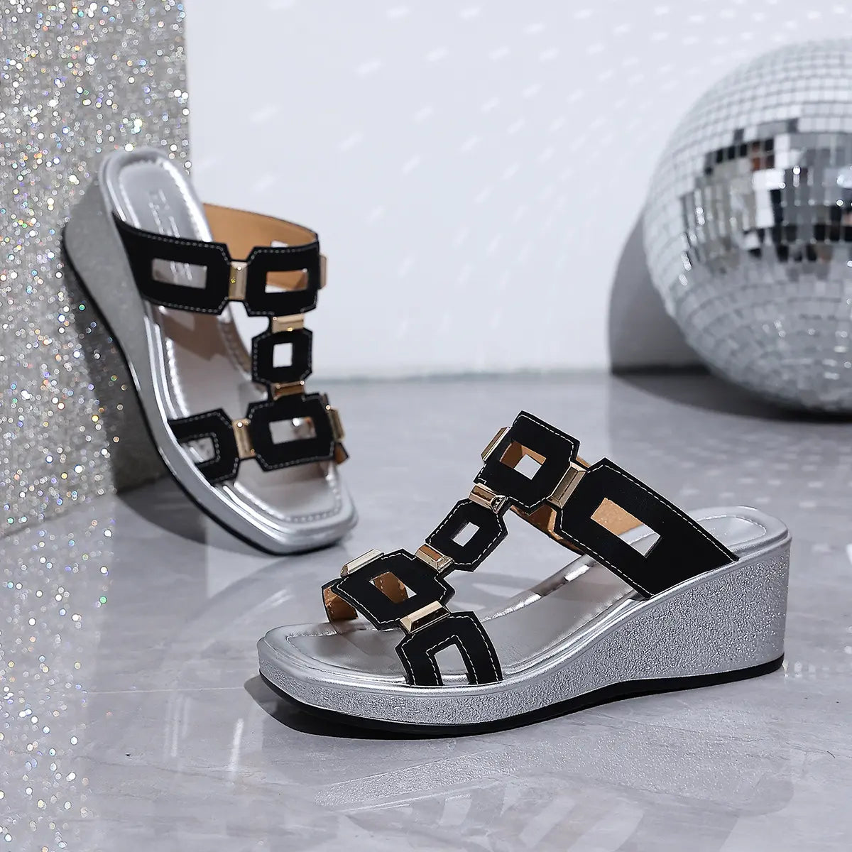 Eskari women's black slip-on wedge sandals with gold geometric hardware, featuring a textured silver wedge and open fish-mouth toe. Presented in a studio setting with a disco ball.