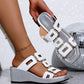 A woman's foot wearing a white Eskari platform wedge sandal with decorative gold geometric hardware and a textured silver wedge heel. A hand holds the matching sandal in a studio setting with a blurred disco ball.