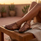 Kevroz chestnut brown woven vegan leather fisherman sandals with gold buckle, on model's feet relaxing on a wooden bench at sunset.