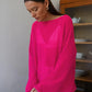 Woman wearing a vibrant fuchsia sheer knit sweater with a relaxed fit, wide boat neckline, and flowing bell sleeves, styled in a bright indoor setting with a vintage cabinet.