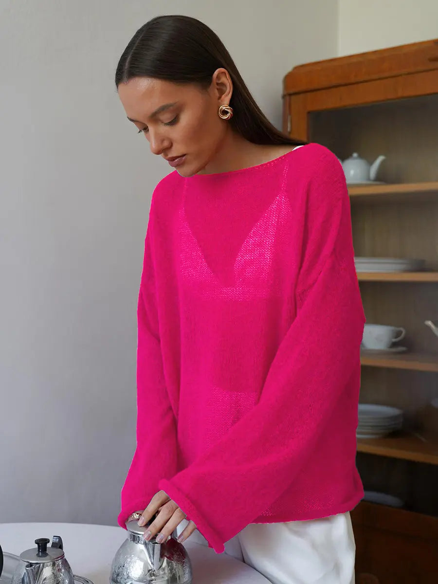 Woman wearing a vibrant fuchsia sheer knit sweater with a relaxed fit, wide boat neckline, and flowing bell sleeves, styled in a bright indoor setting with a vintage cabinet.