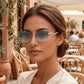 Woman wearing Galyth cat-eye sunglasses with gold frames, crystal-embellished temples, and azure dawn gradient lenses, seated at an outdoor cafe.