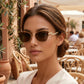 Elegant woman wearing Galyth cat-eye sunglasses with a gold frame, brown lenses, and crystal-embellished temples at an outdoor cafe.