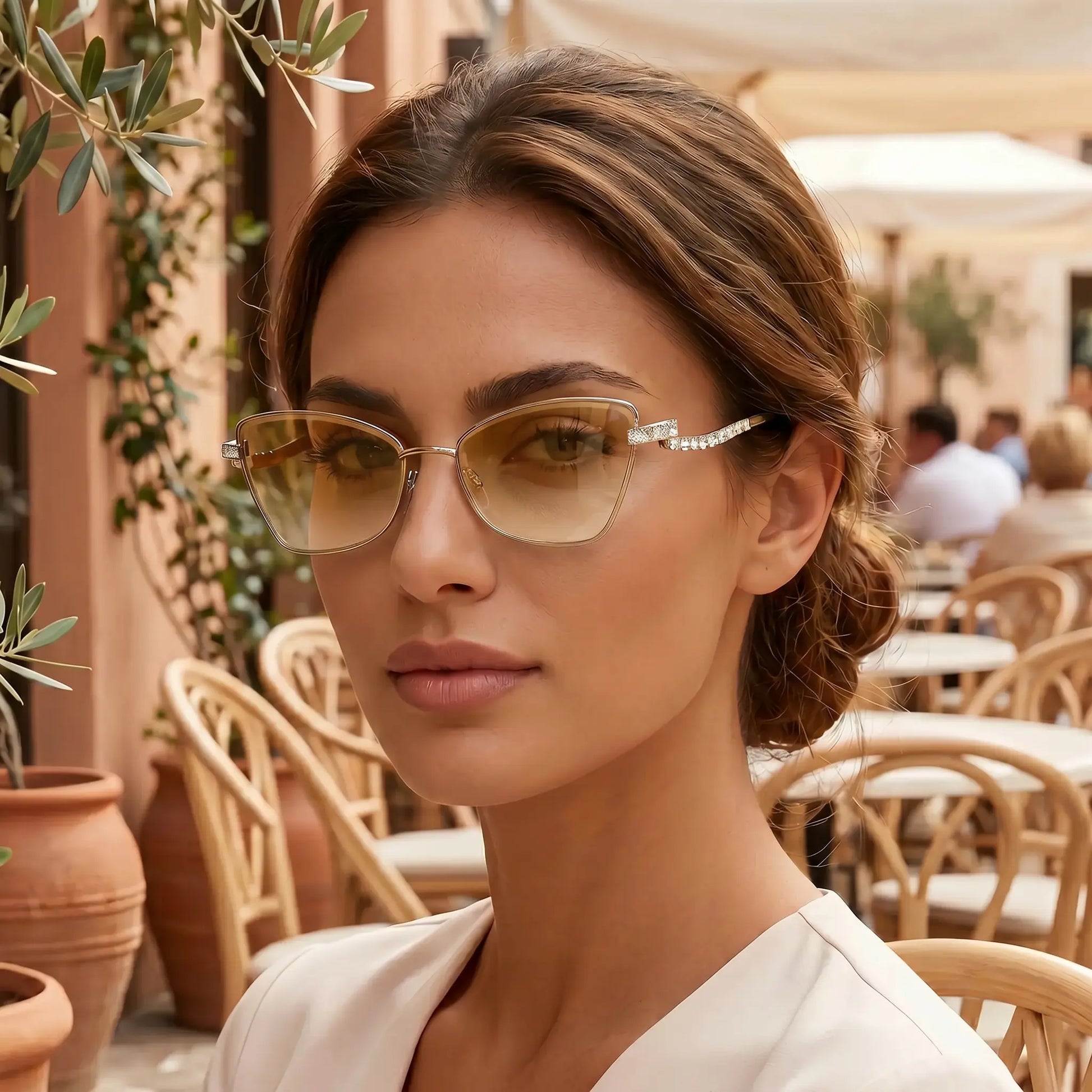Woman wearing Galyth cat-eye sunglasses with gold frames and gradient amber lenses, featuring crystal-embellished zigzag temples, in an outdoor cafe setting.