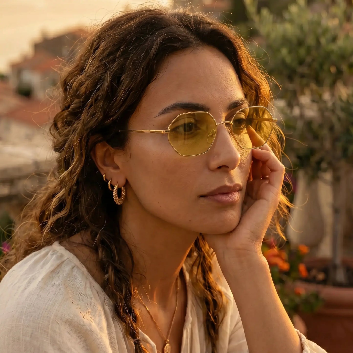 A woman with curly brown hair wears elegant gold-tone geometric octagonal sunglasses with amber tinted lenses, positioned outdoors during golden hour.