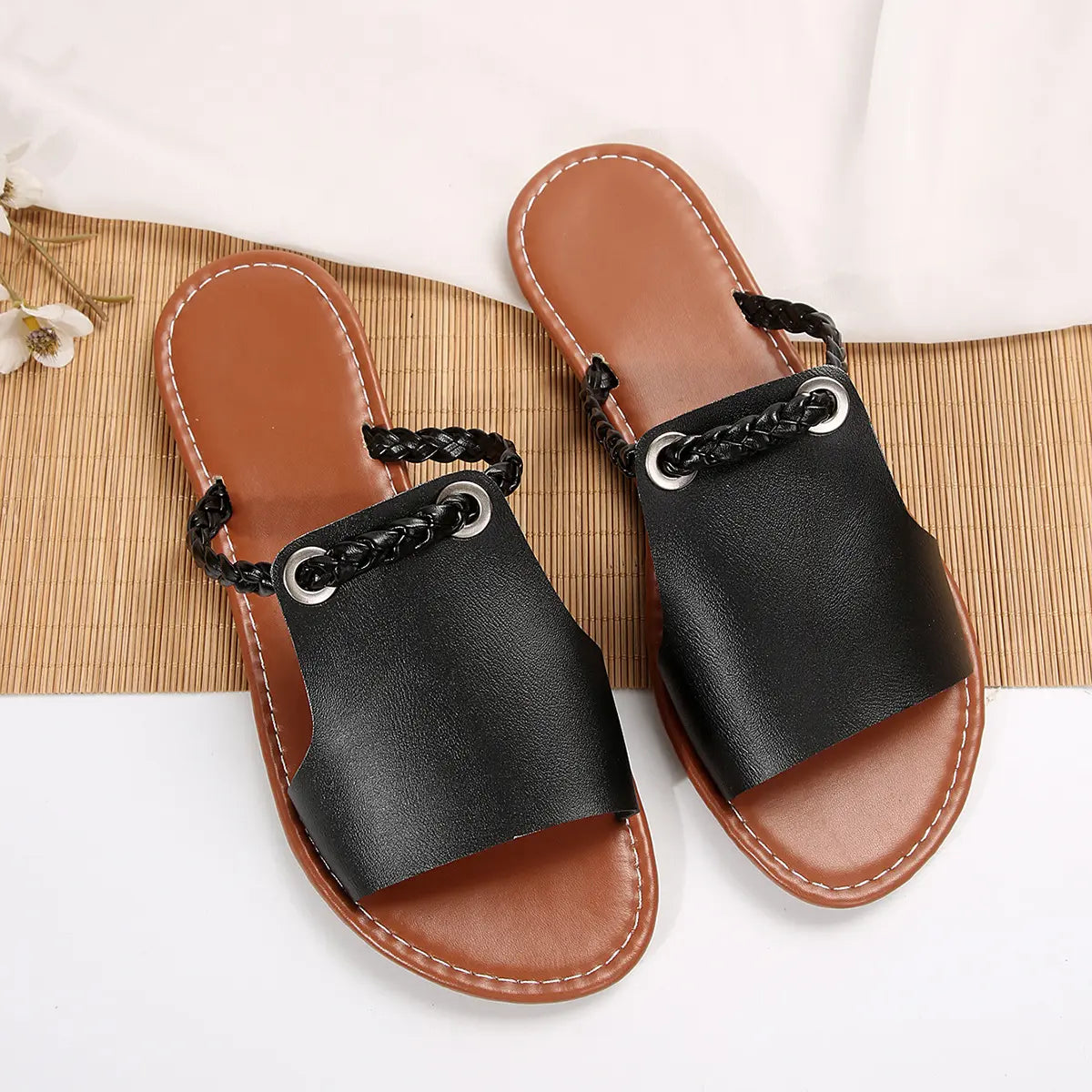 Geylan black flat slide sandals featuring a braided faux leather strap with a polished silver O-ring detail, presented in a studio setting on a textured mat.