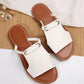 A pair of Geylan flat slide sandals in white faux leather with braided straps and silver O-ring detail, shown from a top-down angle on a bamboo mat with decorative white flowers.