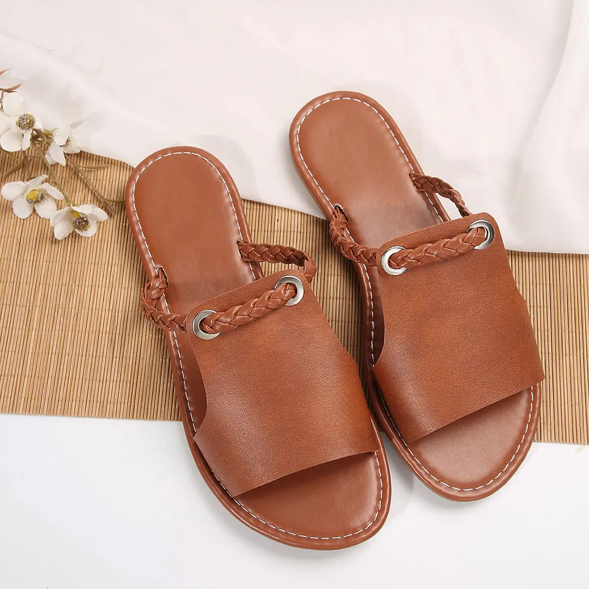 A pair of Geylan flat slide sandals in earthy tan faux leather, featuring a detailed braided strap with a polished silver O-ring accent. These open-toe summer slides are presented on a textured bamboo mat with a draped white cloth and floral accent.
