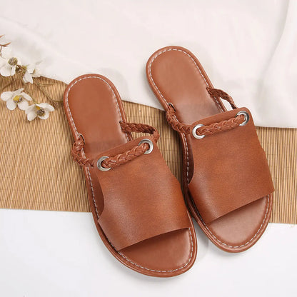 A pair of Geylan flat slide sandals in earthy tan faux leather, featuring a detailed braided strap with a polished silver O-ring accent. These open-toe summer slides are presented on a textured bamboo mat with a draped white cloth and floral accent.