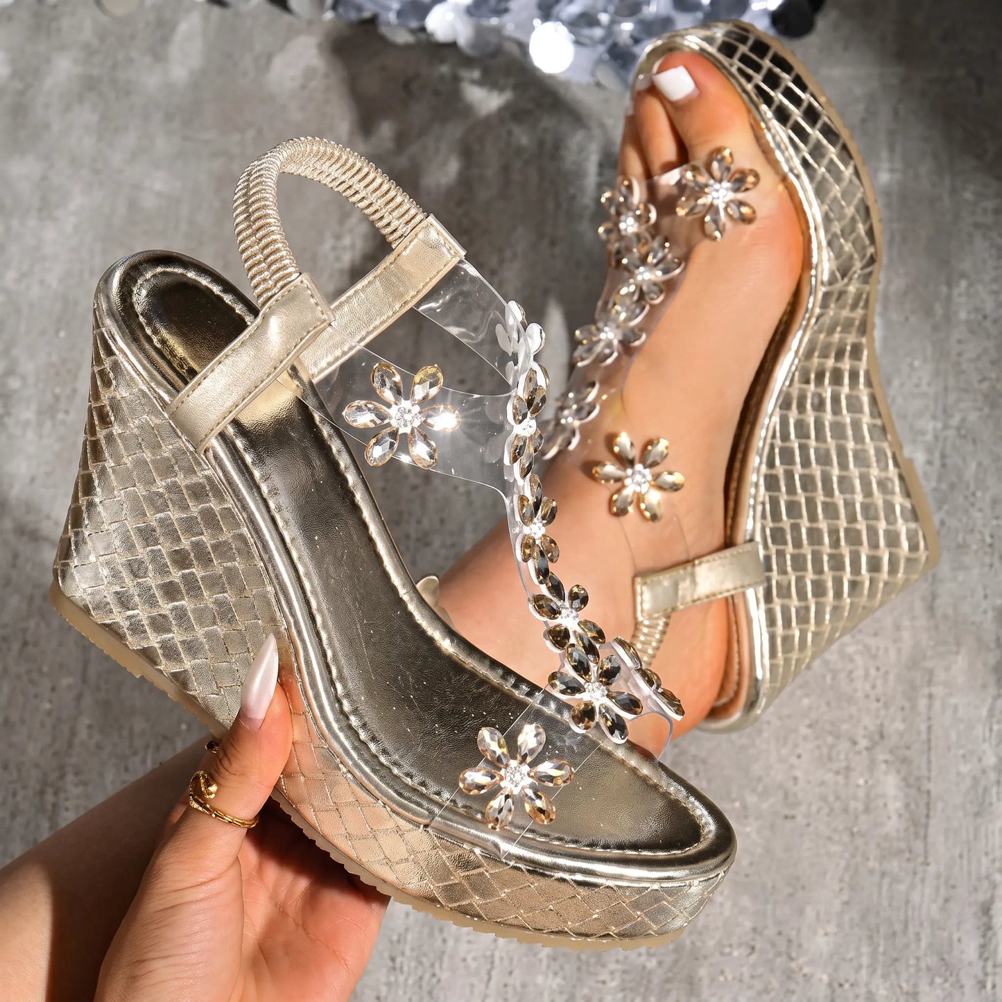 Close-up of a gold women's wedge sandal featuring transparent straps adorned with sparkling crystal floral embellishments and a textured metallic woven wedge heel with a platform sole. A hand holds the heel, showcasing artisanal detail.
