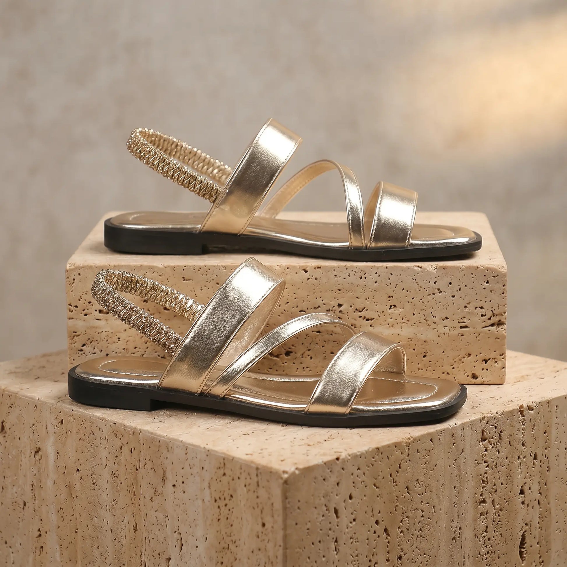Gold Zyneth flat sandals with asymmetrical vegan leather straps, gathered elastic heel, and square toe, displayed on textured blocks.