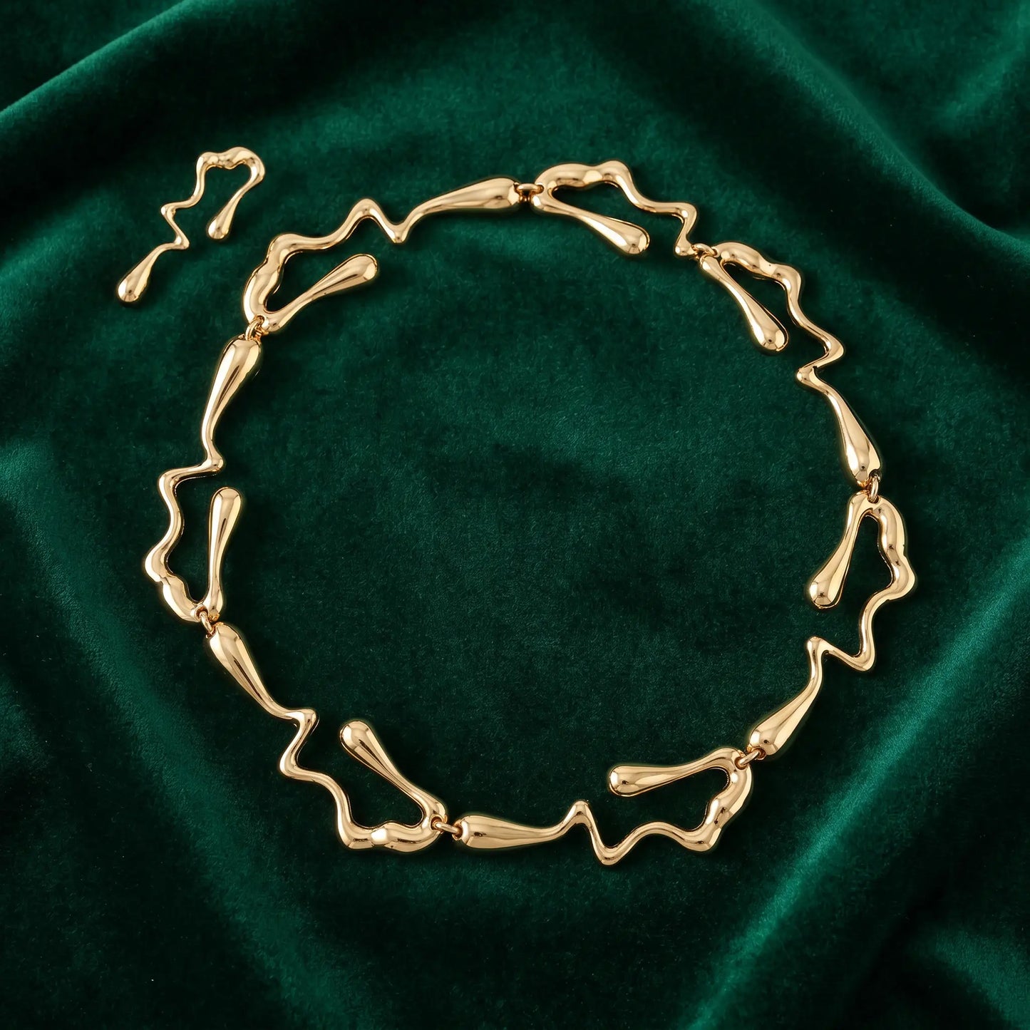 Radiant gold liquid metal choker necklace with abstract wavy links and a coordinating drop earring, displayed on plush emerald velvet.