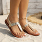 Close-up of women's gold metallic textured flat sandals worn on feet with white toenails, set on a sandy beach with a straw mat. Features thong strap and adjustable ankle strap.