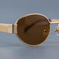 Close-up of gold oval metal frame sunglasses, showing the brown tinted lens, sleek rim, flat hinge detail, and clear adjustable silicone nose pad.
