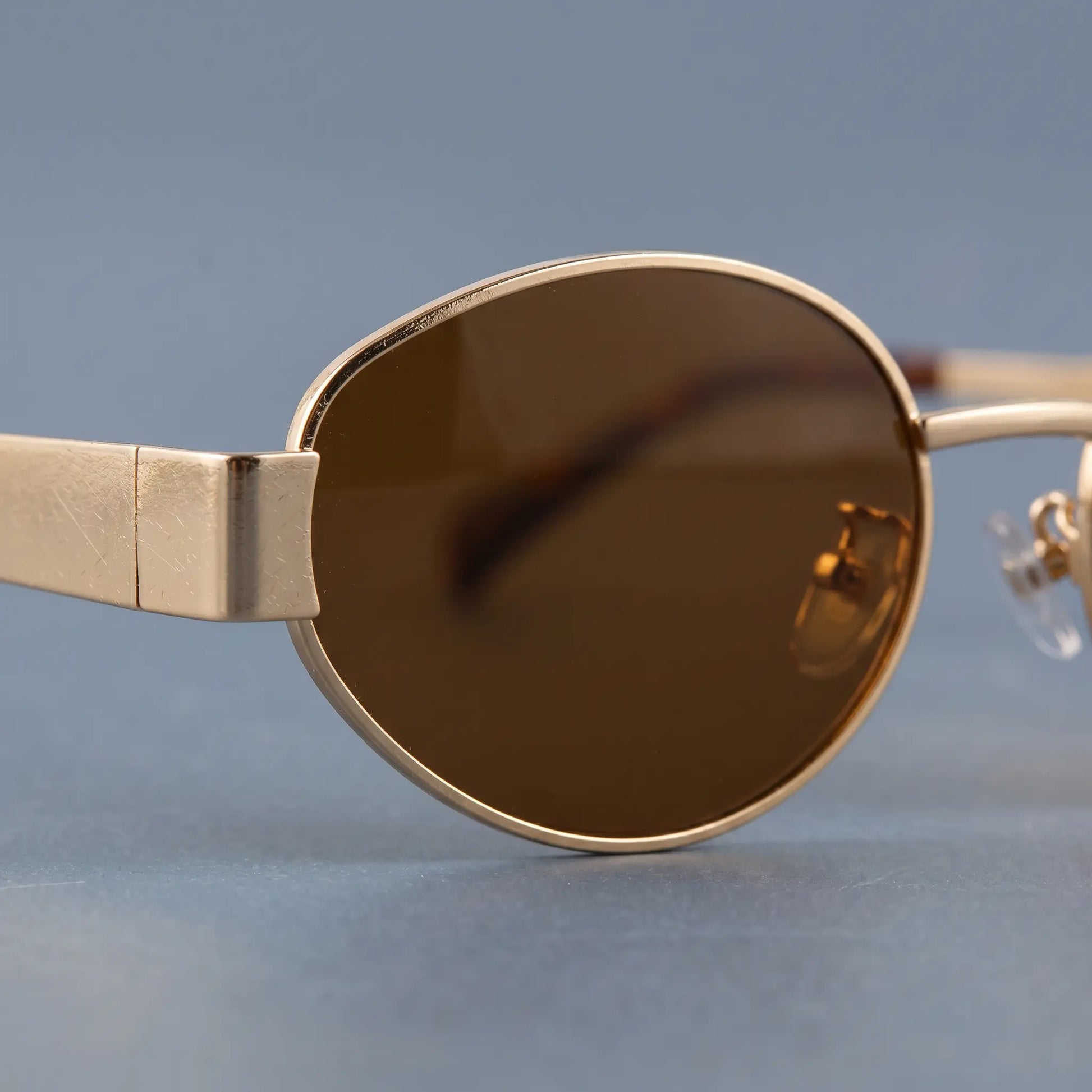Close-up of gold oval metal frame sunglasses, showing the brown tinted lens, sleek rim, flat hinge detail, and clear adjustable silicone nose pad.