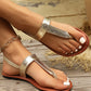 Close-up of women's gold metallic textured flat thong sandals with a T-strap and adjustable ankle buckle, worn on feet with white-painted toenails against a rustic wooden background.