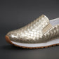 Close-up of a metallic gold slip-on loafer showcasing its intricate woven texture and flat rubber sole. The shoe features a round toe and a comfortable elastic side panel.