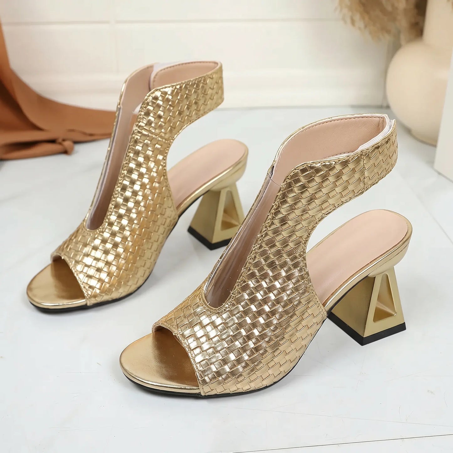 Polished gold peep-toe sandals featuring a woven textured upper, distinctive geometric block heel, and delicate metallic chain ankle strap, displayed in a three-quarter view.