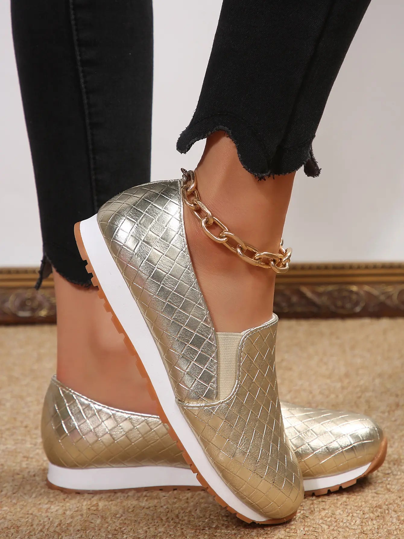 Metallic gold woven slip-on loafers with a round toe, worn with black jeans and a gold anklet, shown from a three-quarter angle.