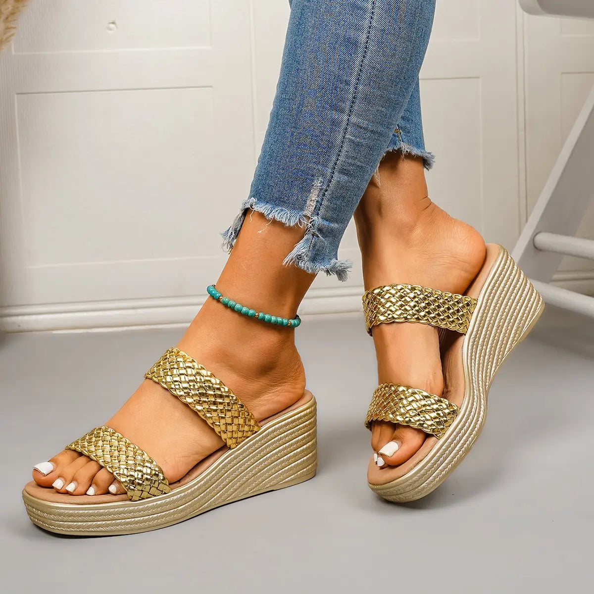 Close-up of a woman wearing gold woven strap platform wedge sandals with a champagne gold sole and tan footbed, paired with distressed blue jeans.