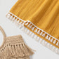 Close-up of the Zovith Golden Sun maxi dress hem, showcasing the vibrant golden-yellow fabric with subtle vertical pointelle stripes and the continuous cream-white tassel fringe trim. A woven straw bag with tassels is partially visible.