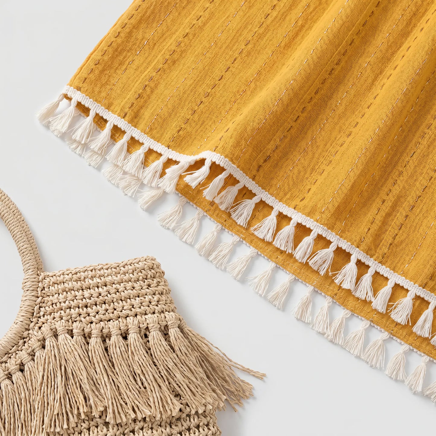 Close-up of the Zovith Golden Sun maxi dress hem, showcasing the vibrant golden-yellow fabric with subtle vertical pointelle stripes and the continuous cream-white tassel fringe trim. A woven straw bag with tassels is partially visible.