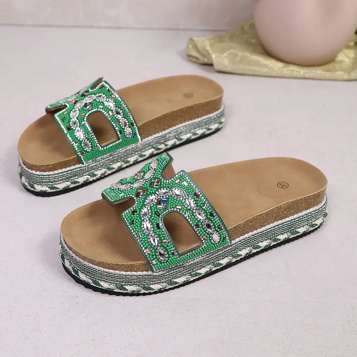 Green embellished platform slide sandals featuring intricate bead and rhinestone detailing on the strap, U-shaped cutouts, a natural cork footbed, and a decorative woven midsole. These open-toe slides offer a bohemian and chic summer style.