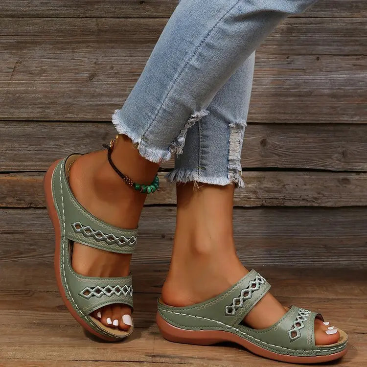 Women's green slip-on slide sandals with geometric embroidered straps, shown on a person's feet with distressed jeans and a wooden background.