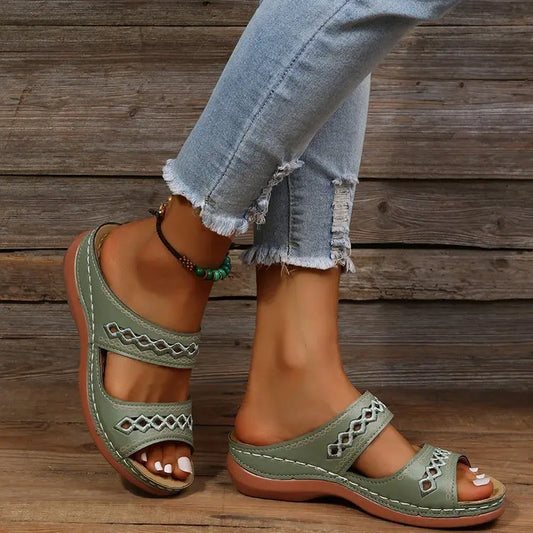 Women's green slip-on slide sandals with geometric embroidered straps, shown on a person's feet with distressed jeans and a wooden background.