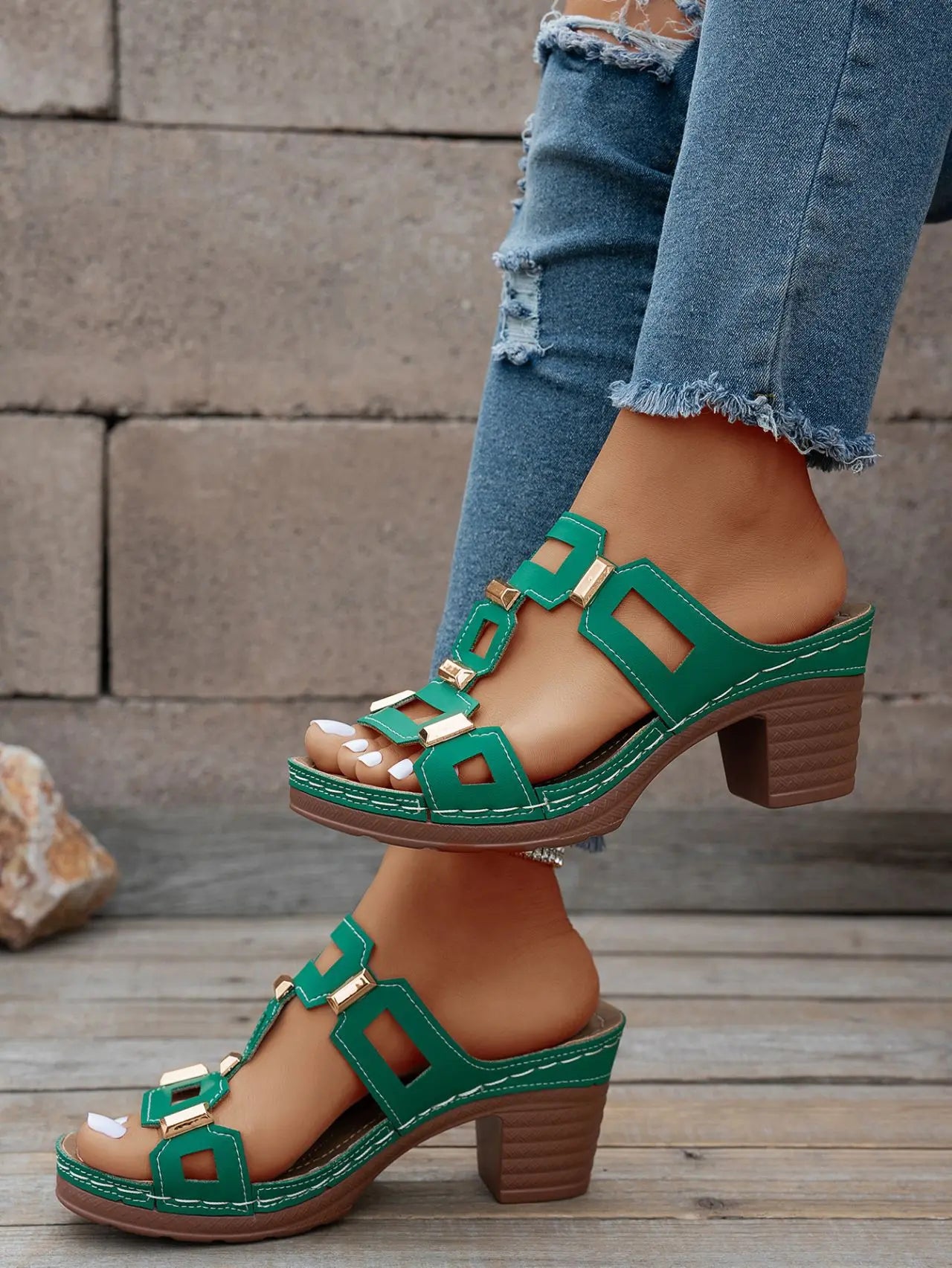 Green heeled platform slide sandals with geometric gold-tone hardware and white contrast stitching, worn with distressed denim jeans.