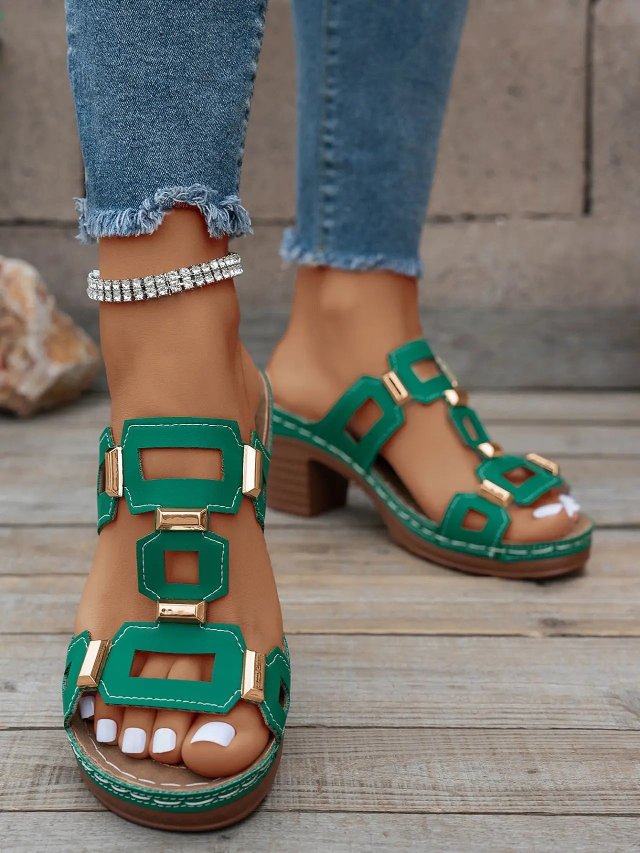 Green heeled platform slide sandals featuring gold-tone geometric hardware and white contrast stitching, worn with distressed jeans and a silver anklet.