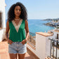 A woman wears a sleeveless emerald green blouse with a decorative cream crochet V-neck trim, paired with denim shorts, standing on a sun-drenched balcony overlooking the Mediterranean Sea and white-washed village.