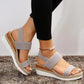 Women's grey espadrille wedge sandals featuring elasticated straps, a peep-toe design, and a decorative gold-tone ankle chain, shown on feet with black trousers.