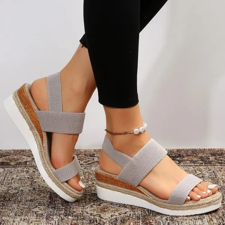 Women's grey espadrille wedge sandals featuring elasticated straps, a peep-toe design, and a decorative gold-tone ankle chain, shown on feet with black trousers.
