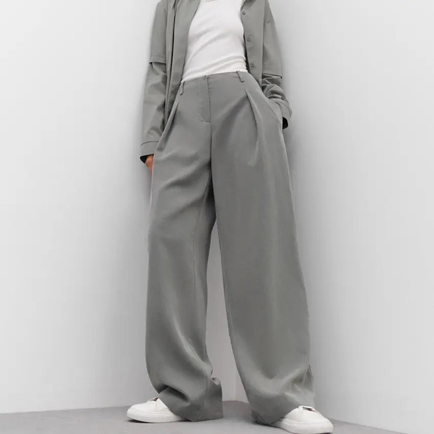 Sophisticated high-waisted wide-leg trousers in a muted grey, featuring sharp front pleats and a fluid drape, paired with a white top and sneakers.