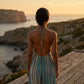 Back view of a woman wearing a Zendru turquoise tie-dye halter maxi resort dress with a deep open back, overlooking a coastal sunset.